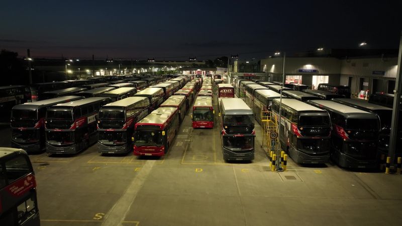A fleet of red double-decker buses parked in a large depot at night, illuminated by streetlights, conveying a calm and orderly atmosphere.