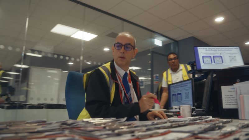 Office setting with two men in high-visibility vests. One is seated at a desk cluttered with documents, looking focused. A colleague stands behind him.