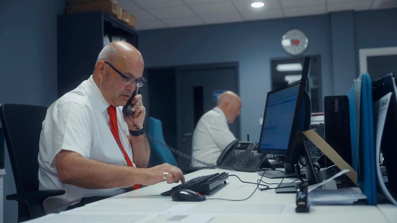 A man in a white shirt with a red tie works at an office desk, talking on the phone and using a computer. Another person is visible in the background.