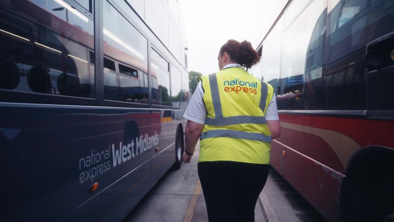 A person in a high-visibility vest walks between two buses at a depot. The vest reads "national express". The scene feels businesslike and focused.