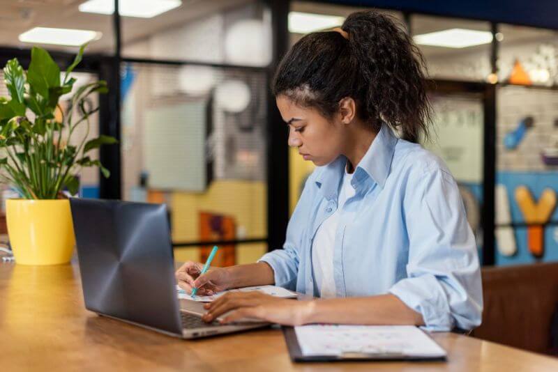 A woman in a blue shirt focuses intently on writing at a wooden table. A laptop is open beside her, and a potted plant adds a touch of greenery.